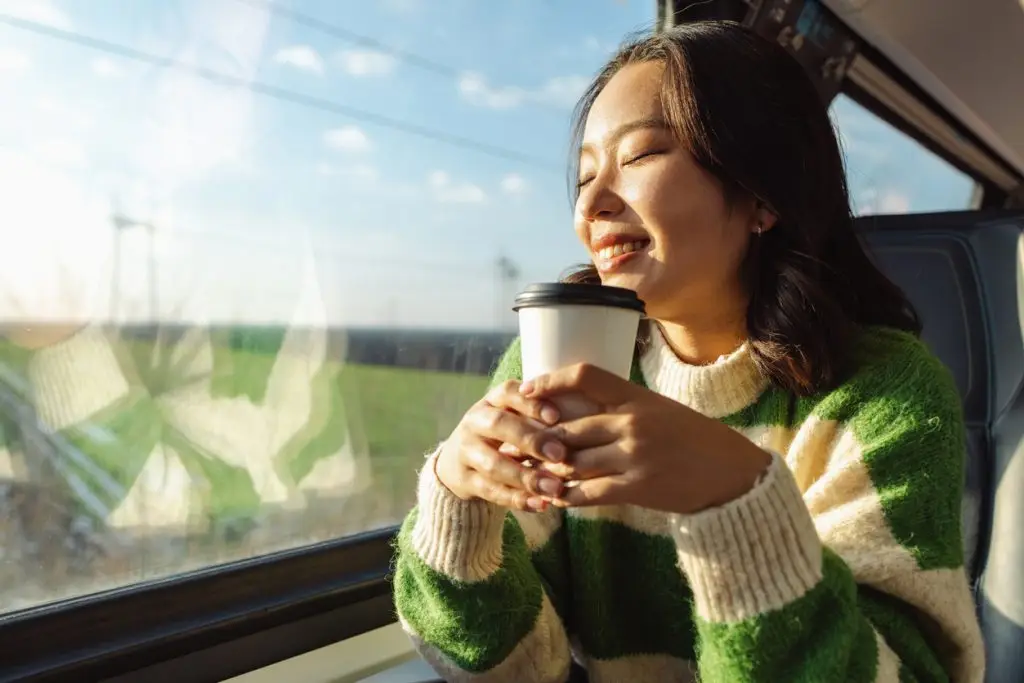 A cheerful young woman smiles as she enjoys a relaxing train journey, holding a warm cup of coffee. The view outside shows a sunny day, enhancing the tranquil and joyful experience.
