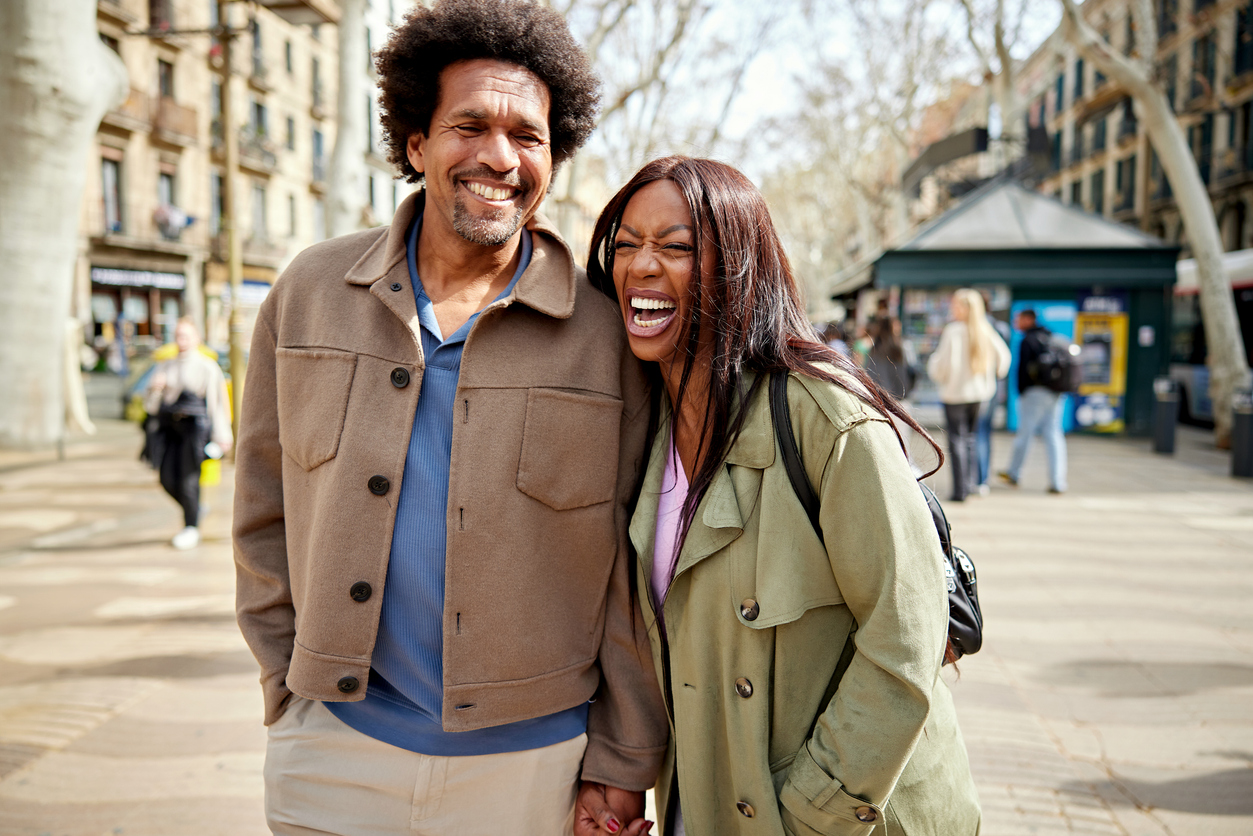 Waist up view of casually dressed man and woman in 40s and 50s, walking outdoors, talking, smiling, and laughing.