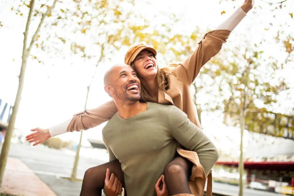 Happy couple enjoying outdoors with piggyback ride, laughing and arms outstretched